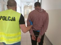 obraz przedstawia policjanta w żółtej kamizelce z napisem Policja, który prowadzi zatrzymanego mężczyznę
